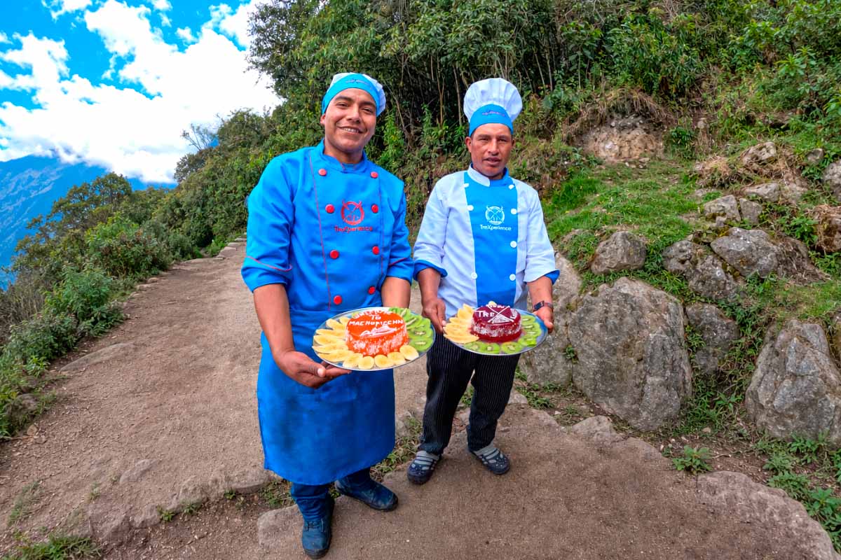 Cocineros con uniforme azul sosteniendo platos con comida en un sendero de montaña rodeado de vegetación | TreXperience