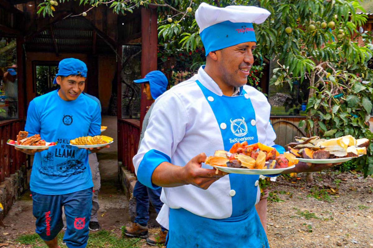 Cooks with food dishes outdoors Chefs in blue uniforms holding plates of food served outdoors, surrounded by greenery | TreXperience