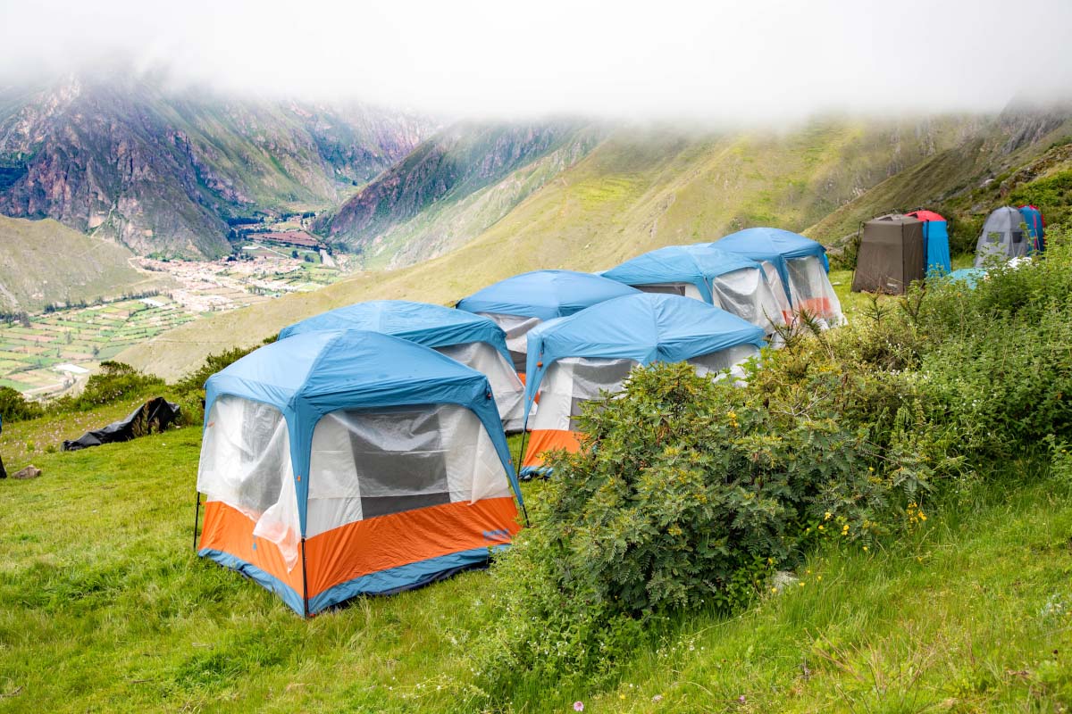 Camp on a green hillside between mountains Several tents set up on a green hillside, with mountains and low clouds in the background | TreXperience