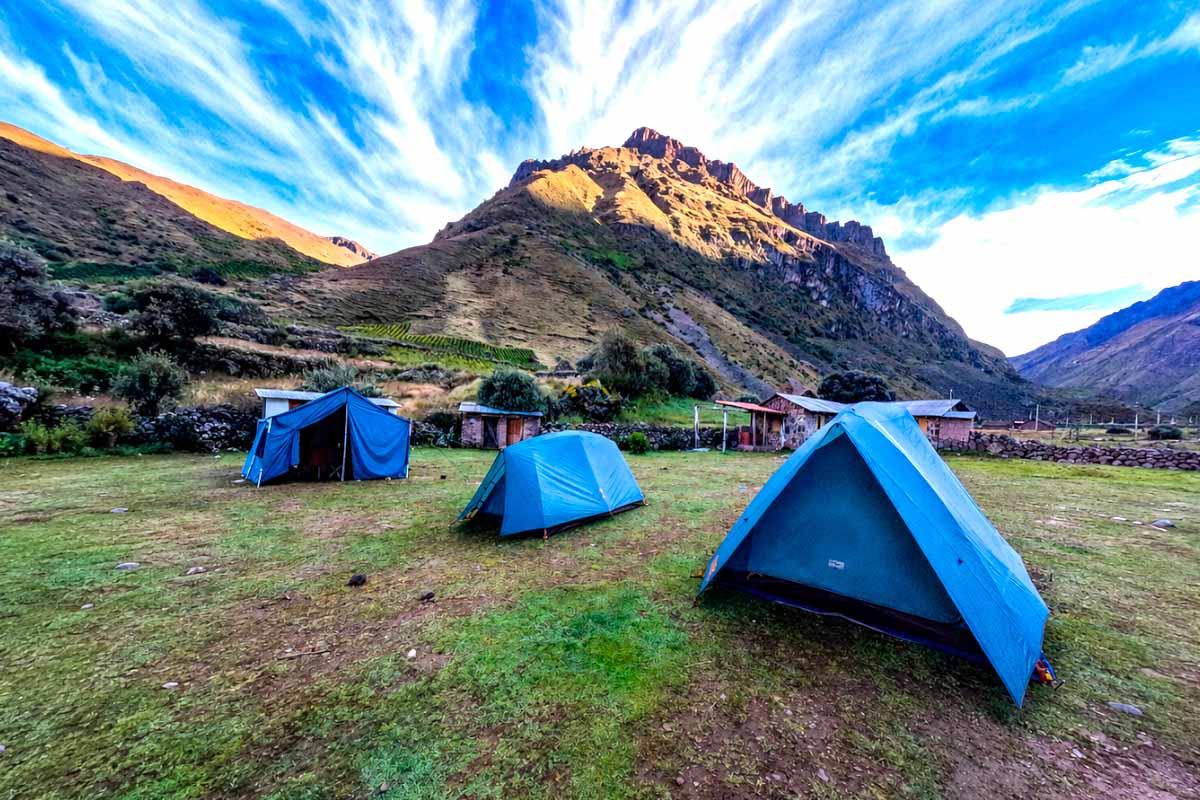 Tiendas de campaña azules instaladas sobre una zona verde frente a una montaña rocosa bajo un cielo con nubes | TreXperience