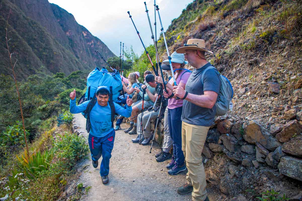 Grupo de porteadoresexcursionistas caminando por un sendero estrecho de montaña entre vegetación y una pared rocosa | TreXperience