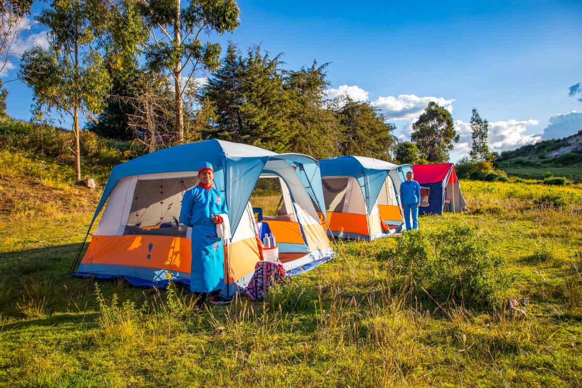 Varias tiendas de campaña azules y naranjas instaladas sobre una zona verde, con árboles y montañas al fondo | TreXperience