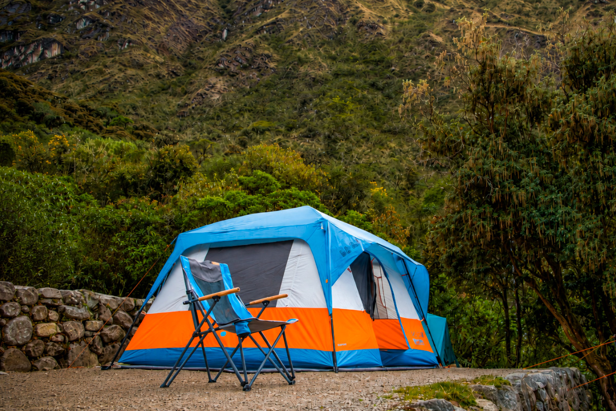 Tienda de campaña azul y naranja instalada junto a un muro de piedra, rodeada de vegetación y montañas | TreXperience