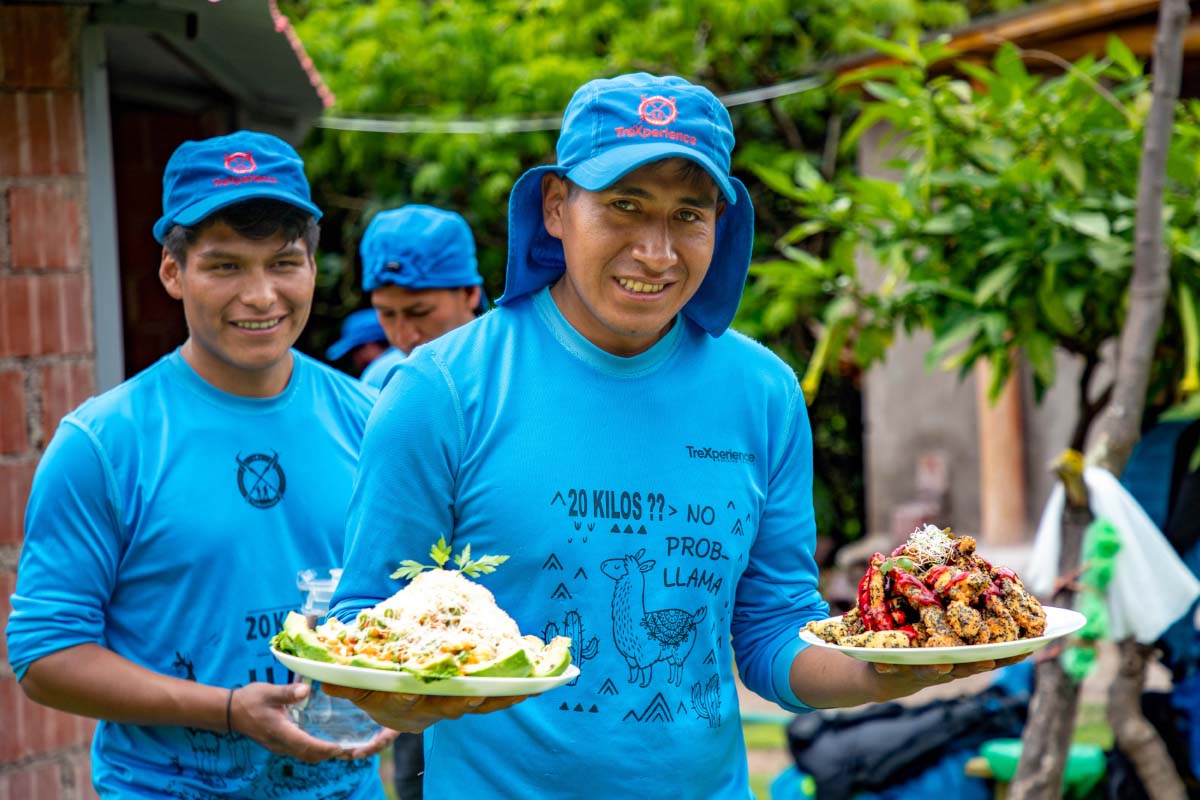 Asistentes de Cocina con uniforme azul sosteniendo platos con comida servida, rodeadas de vegetación al aire libre | TreXperience