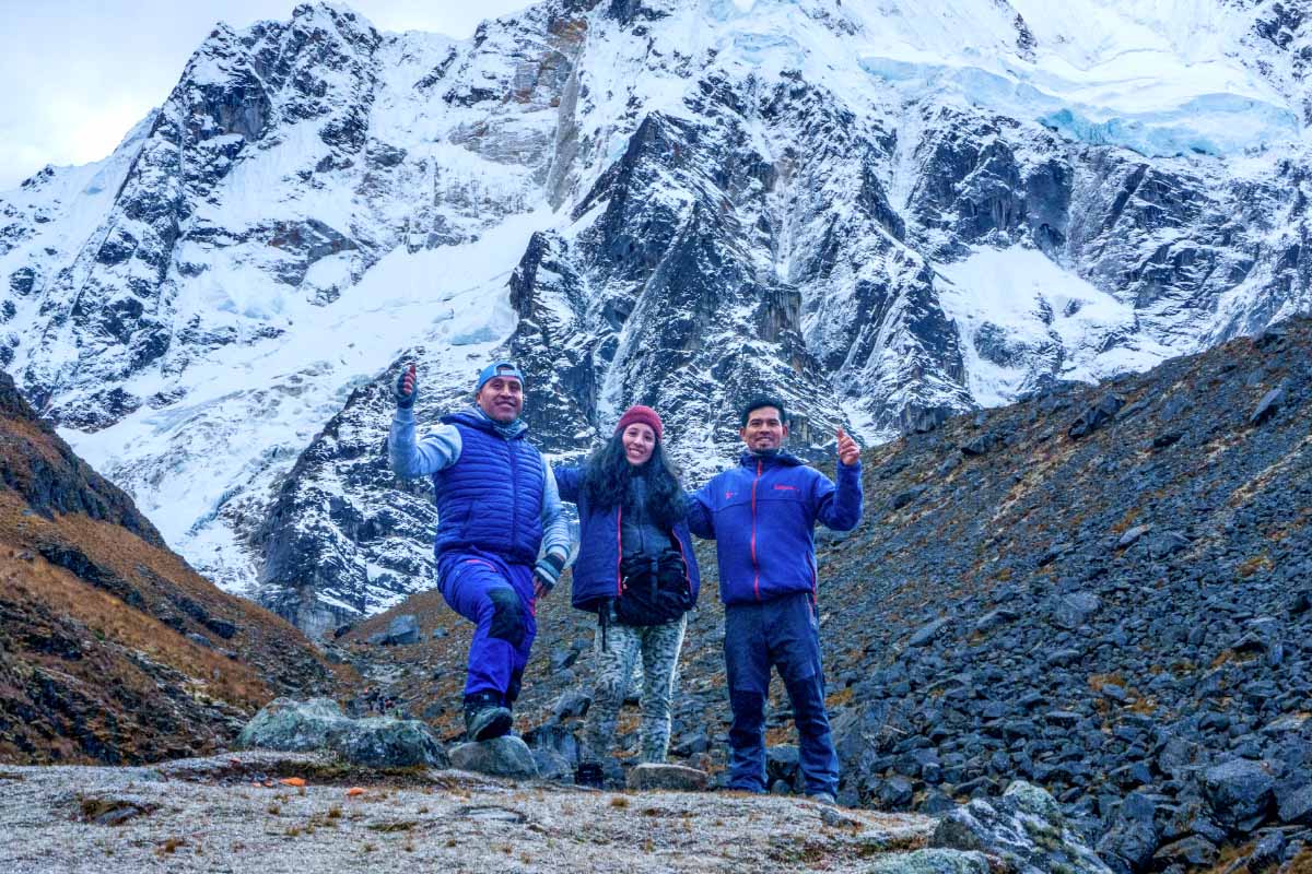 Excursionistas frente a montañas nevadas junto a su guia mujer Dos excursionistas de pie frente a montañas nevadas en un paisaje andino rocoso | TreXperience