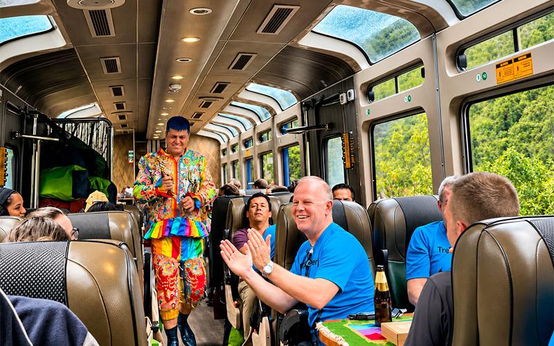 Pasajero aplaudiendo a un bailarín con traje multicolor dentro de un tren panorámico