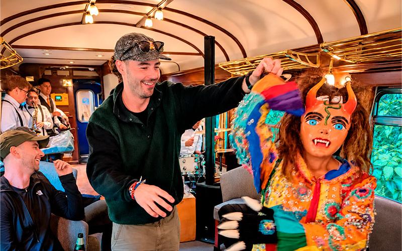 Viajero bailando con un danzante de máscara con cuernos y traje bordado junto a músicos a bordo de un tren