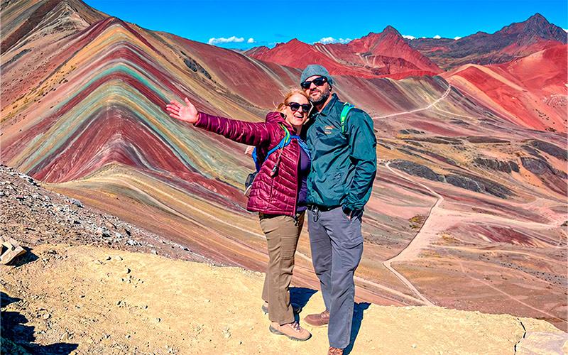 Dos senderistas posando abrazados frente a Vinicunca, la Montaña de Siete Colores, con senderos al fondo