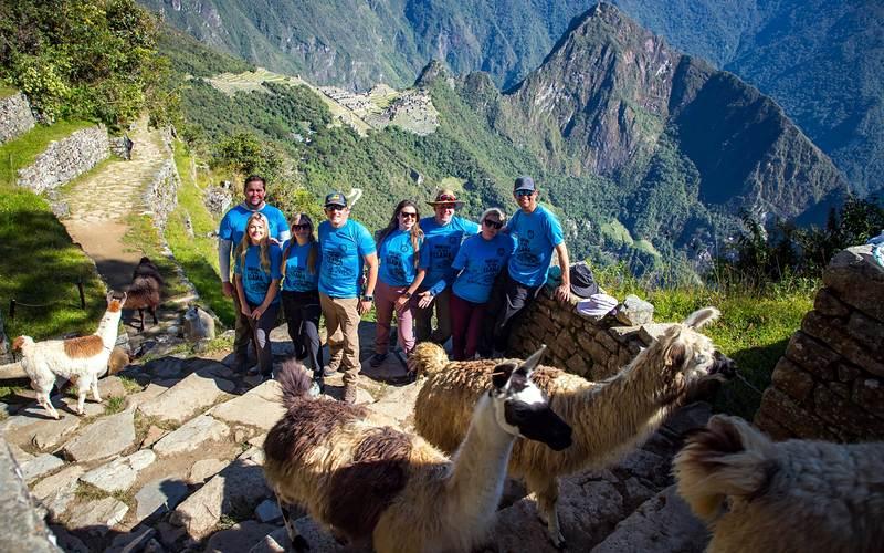 Group of hikers in TreXperience shirts posing with llamas on the Inca Trail, with Machu Picchu below