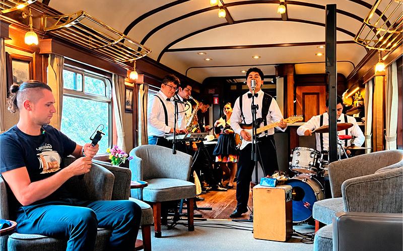Músicos tocando guitarra, saxofón, teclado y batería en el salón de un tren junto a un pasajero sentado