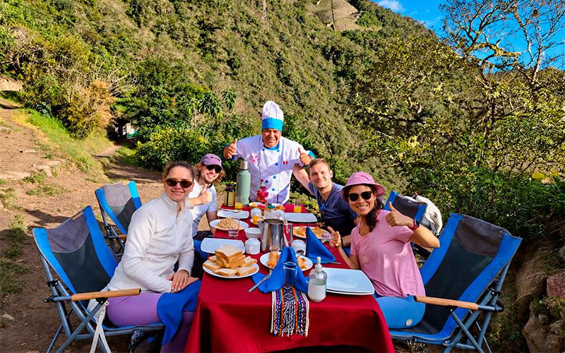 Four hikers and a chef posing around a table set for an outdoor meal, with stone terraces on the hillside