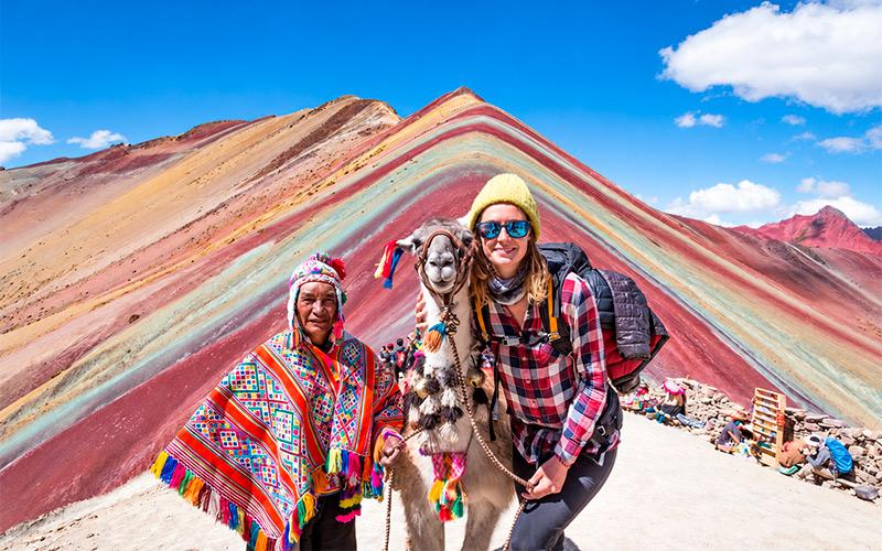 Viajera junto a una llama y un hombre con poncho multicolor frente a Vinicunca, la Montaña de Siete Colores