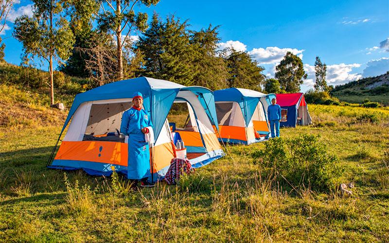 Two staff members in blue uniforms beside glamping tents on a grassy campsite surrounded by trees