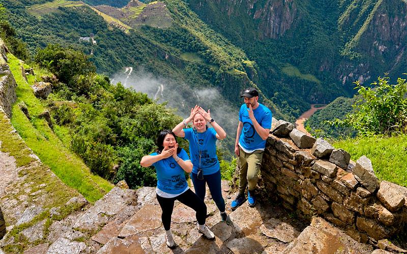 Three travelers in blue shirts posing on the stone steps of Intipunku, with Machu Picchu in the background