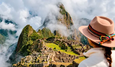 Traveler in a beige hat seen from behind overlooking Machu Picchu, with Huayna Picchu partly covered in mist