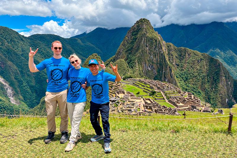 Tres viajeros con camisetas azules de TreXperience frente a Machu Picchu con Huayna Picchu al fondo