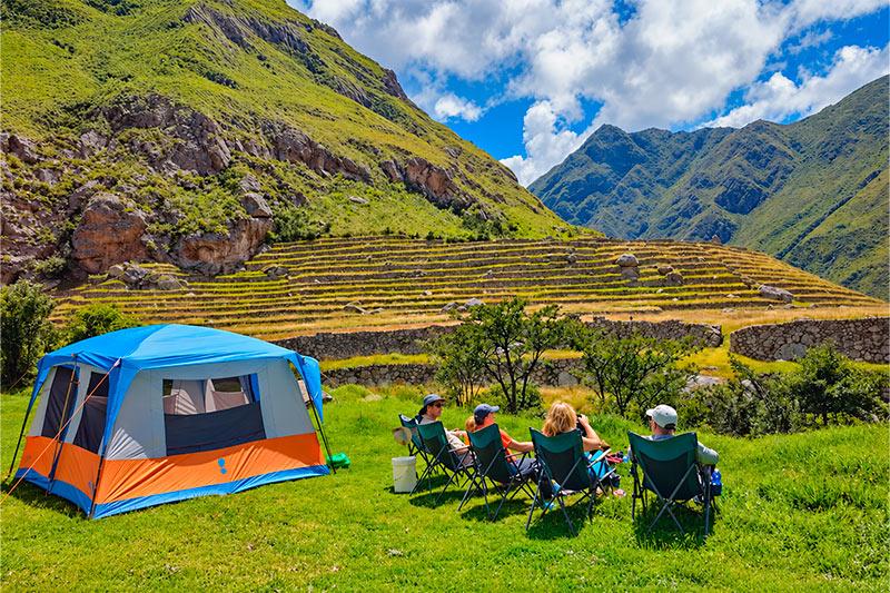 Cuatro senderistas descansando junto a una carpa azul y naranja, frente a andenes de piedra y montañas verdes