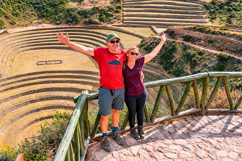 Two travelers posing with their arms outstretched above the circular Inca terraces of Moray