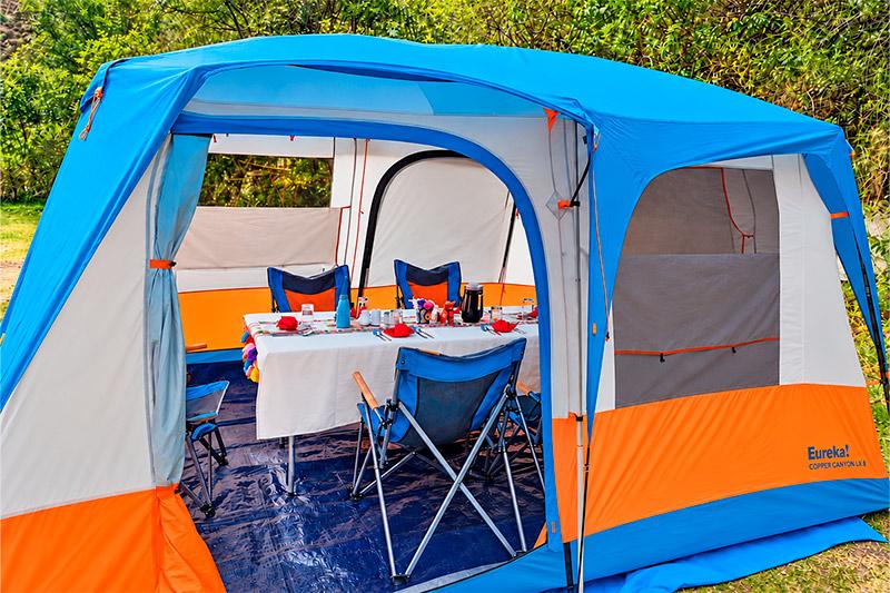 Carpa comedor azul y naranja con mesa puesta, sillas plegables y alfombras estampadas