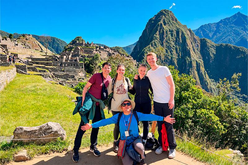Cuatro viajeros y su guía posando en Machu Picchu con construcciones incas y Huayna Picchu al fondo