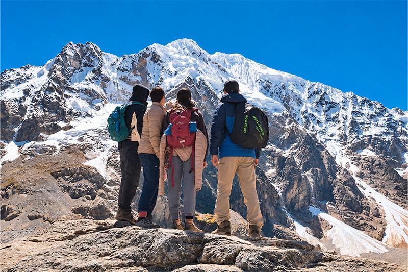 Cuatro senderistas con mochilas en un mirador rocoso, de espaldas frente a una montaña nevada bajo un cielo azul