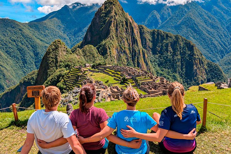 Cuatro viajeros sentados de espaldas y abrazados frente a Machu Picchu con Huayna Picchu al fondo