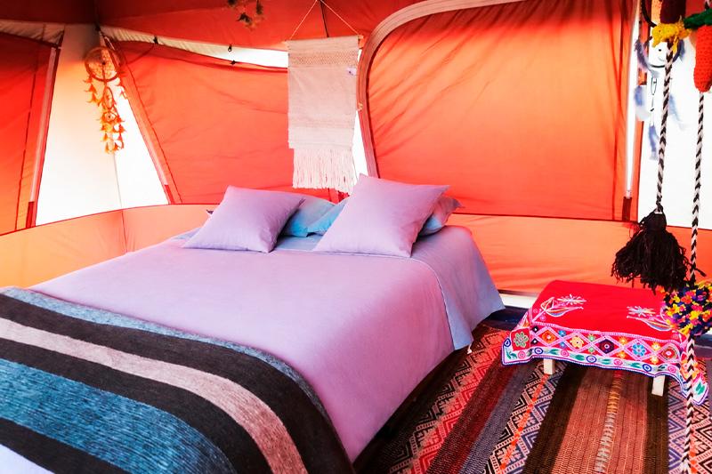 Interior de carpa de glamping con cama doble, ropa de cama lila, alfombras tejidas y mesa con mantel bordado