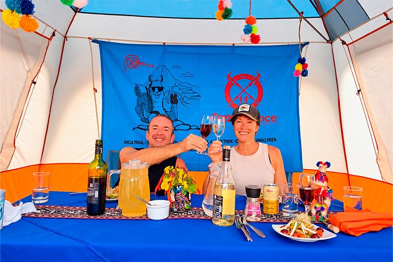 Dos viajeros brindando ante una mesa con comida y bebidas dentro de una carpa comedor de TreXperience