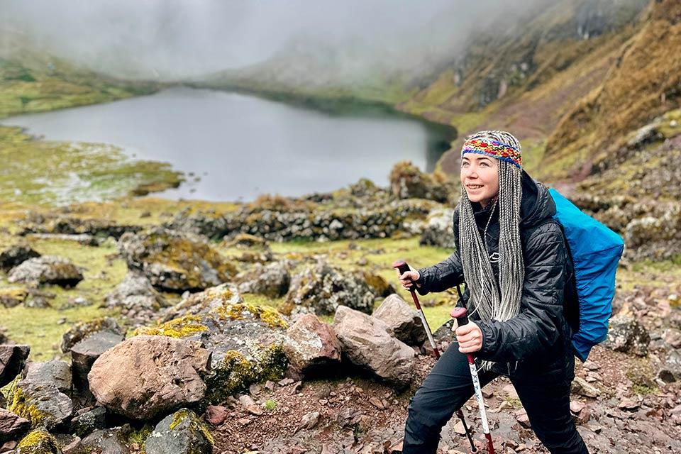 Caminante con bastones y mochila sube por sendero rocoso con una laguna de altura al fondo, Trek Lares y Camino Inca Corto 5 días | TreXperience
