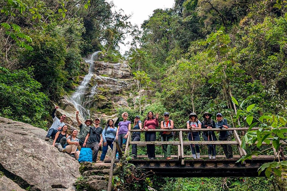 Grupo de turistas posa en un puente de madera frente a una cascada rodeada de vegetación, Trek Lares y Camino Inca Corto 5 días | TreXperience