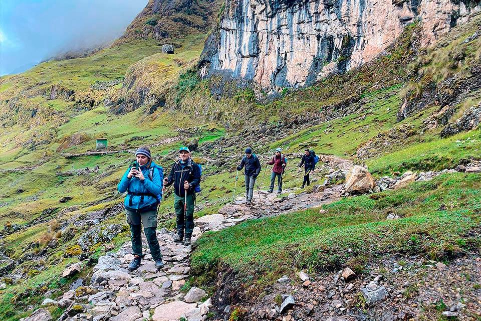 Grupo de caminantes avanza por un camino de piedra junto a una gran pared rocosa en laderas verdes, Trek Lares y Camino Inca Corto 5 días | TreXperience
