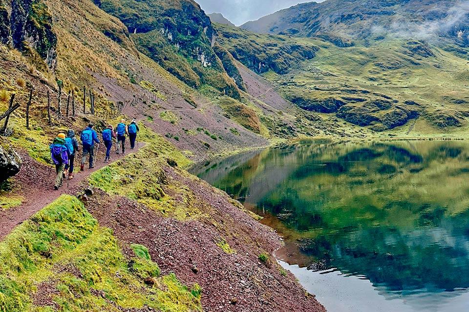 Caminantes con mochilas avanzan por un sendero junto a una laguna verde en valle de montaña, Trek Lares a Machu Picchu 4 días | TreXperience