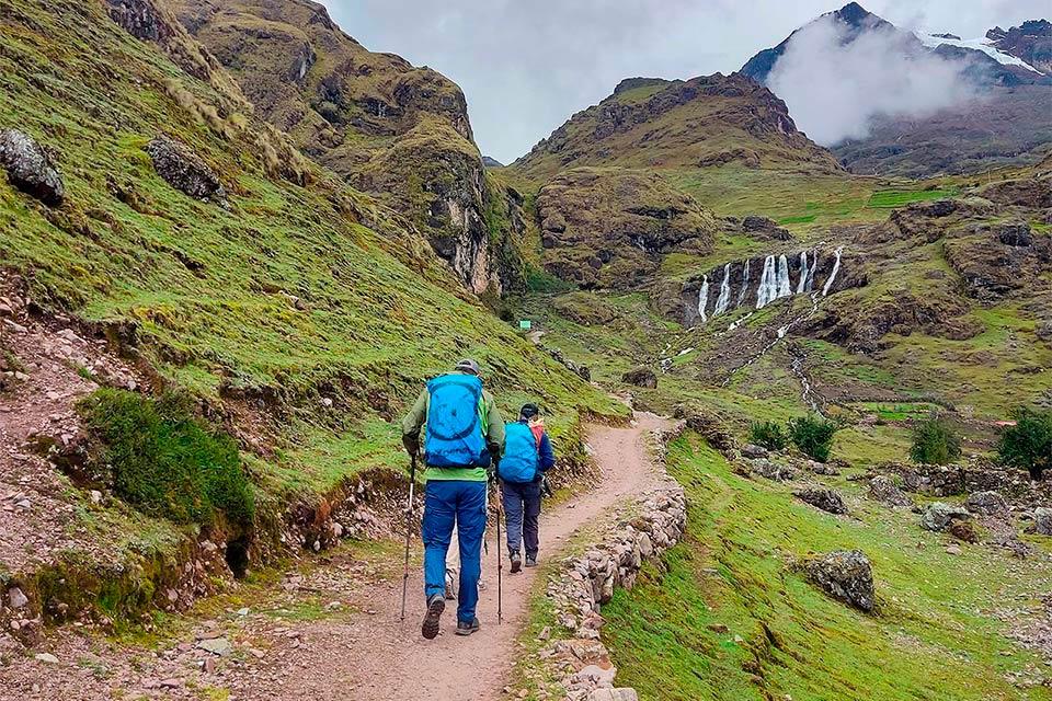 Dos caminantes avanzan por un sendero en valle verde con cascadas al fondo y nubes bajas, Trek Lares a Machu Picchu 4 días | TreXperience