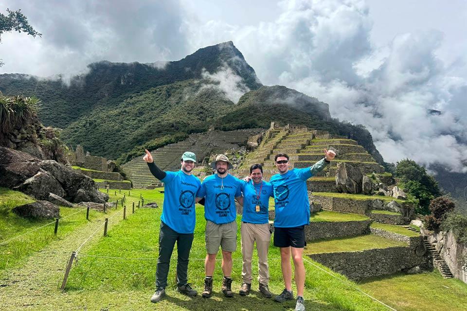 Cuatro visitantes con polos azules posan en Machu Picchu con andenes y montañas con nubes al fondo, Trek Lares a Machu Picchu 4 días | TreXperience