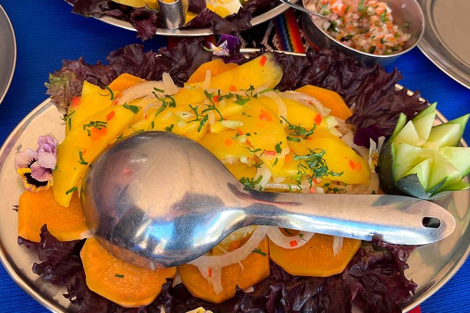 Plato de ceviche de mango con rodajas de camote, cebolla y hierbas sobre hojas moradas, con cucharón metálico encima, Trek Lares a Machu Picchu 4 días | TreXperience