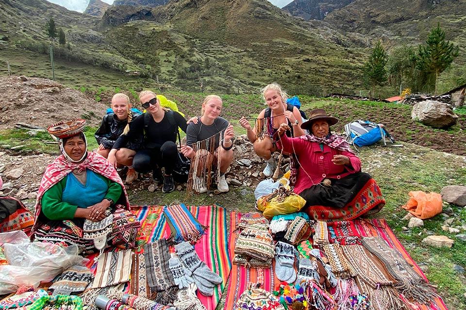 Grupo posa con dos mujeres locales junto a mantas con textiles y artesanías extendidas en el suelo en un valle de montaña, Trek Lares a Machu Picchu 4 días | TreXperience