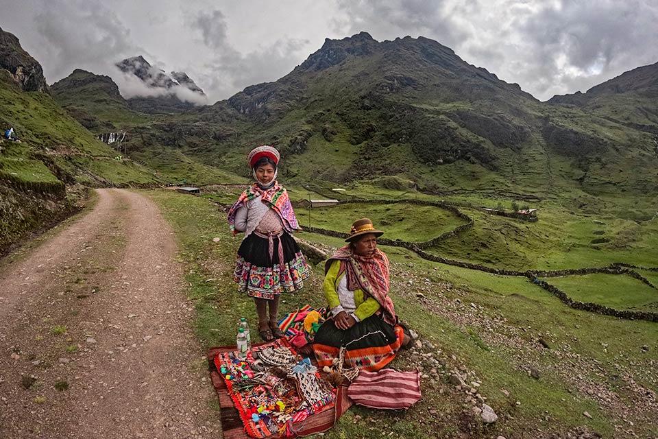 Dos mujeres con ropa tradicional junto a mantas con artesanías al borde de un camino en valle verde de montaña, Trek Lares a Machu Picchu 4 días | TreXperience