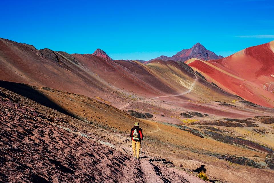 Hiker walking along a trail between red and yellow slopes in the Red Valley under a clear blue sky, Rainbow Mountain and Red Valley Premium Tour | TreXperience