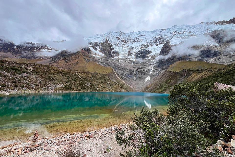 Vista de la laguna Humantay de color turquesa con orilla rocosa y montañas nevadas con nubes bajas al fondo, Caminata Salkantay y Laguna Humantay 2 días | TreXperience