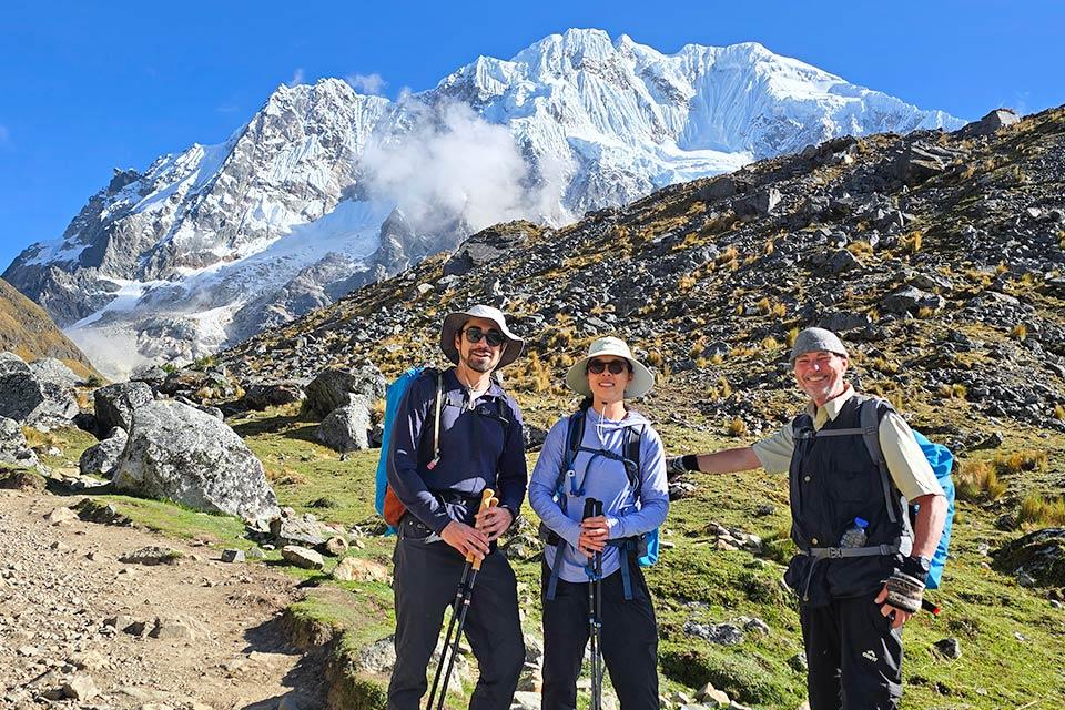 Tres caminantes posan con bastones en un sendero rocoso con una montaña nevada al fondo, Caminata Salkantay y Laguna Humantay 2 días | TreXperience