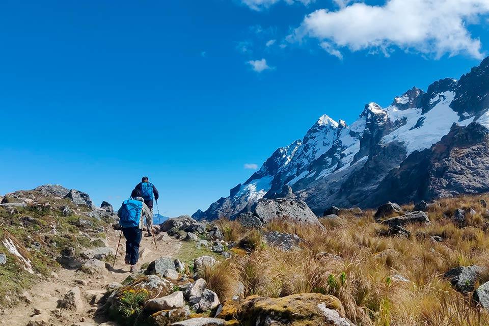 Dos caminantes con mochilas y bastones suben por un sendero de tierra hacia una montaña nevada, Caminata Salkantay y Laguna Humantay 2 días | TreXperience
