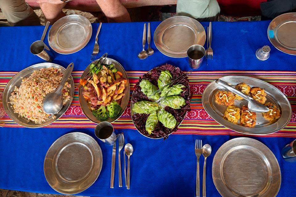 able with a blue tablecloth and an Andean textile runner in the middle, with metal plates, cutlery, and serving dishes of food, Salkantay Trek 4 Days to Machu Picchu | TreXperience