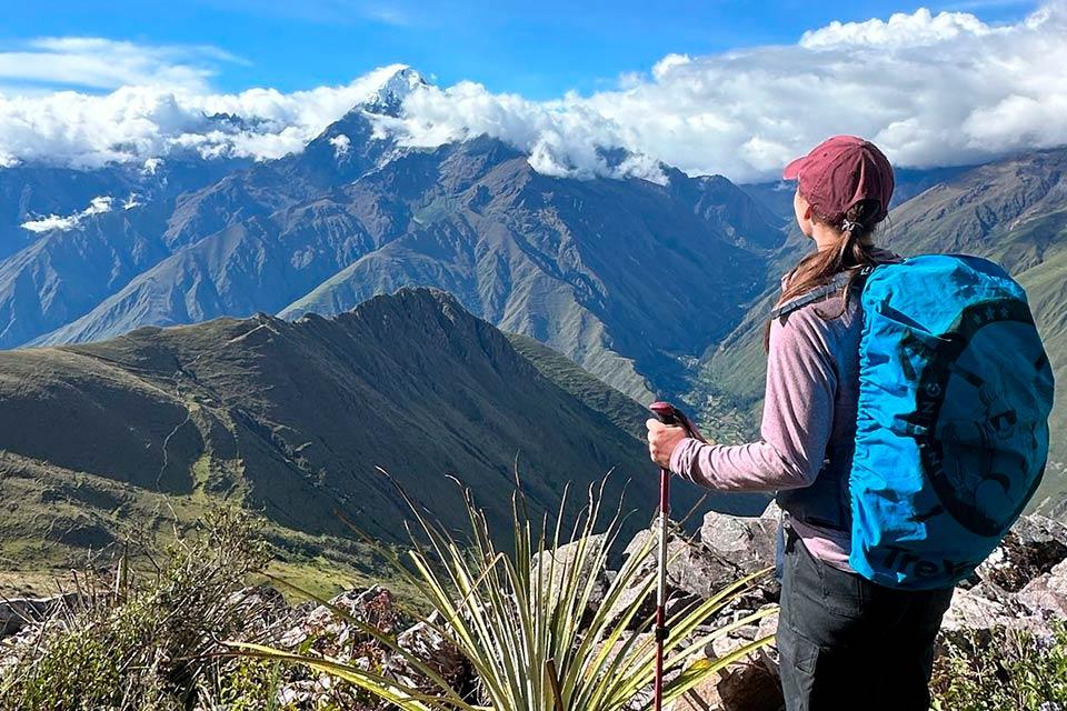 Hiker with a cap and trekking pole looks over a mountain valley from a rocky slope, with a snowcapped peak and clouds behind, Cachicata Inca Quarry Trek to Machu Picchu 4 Days 3 Nights | TreXperience
