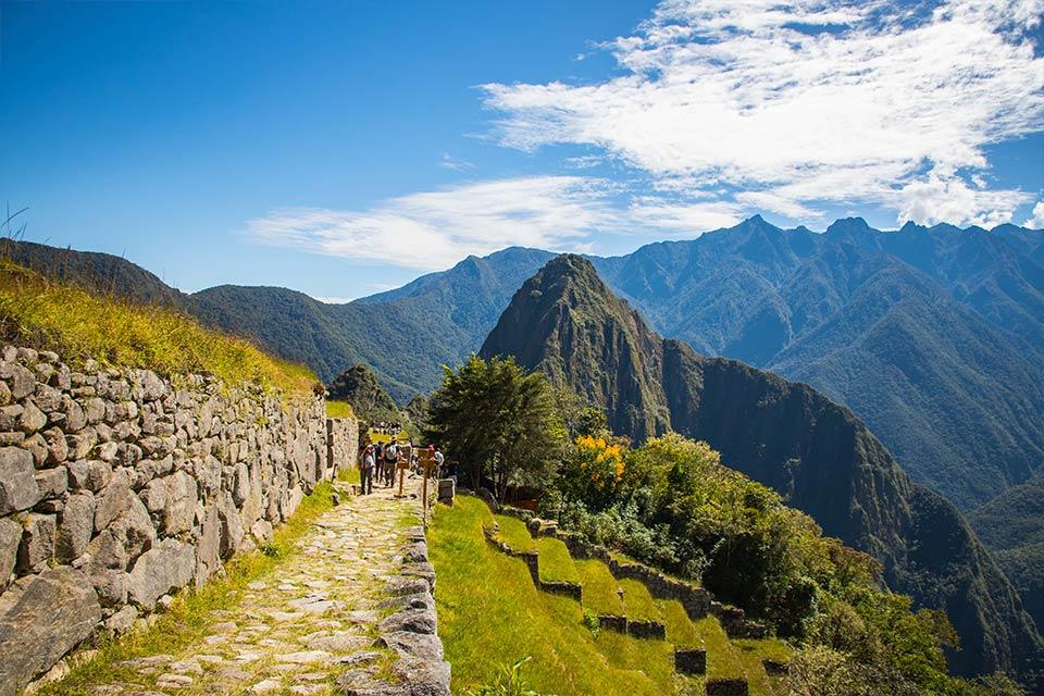 Stone path beside a wall and hillside terraces, with Machu Picchu in the distance and mountains behind, Cachicata Inca Quarry Trek to Machu Picchu 4 Days 3 Nights | TreXperience
