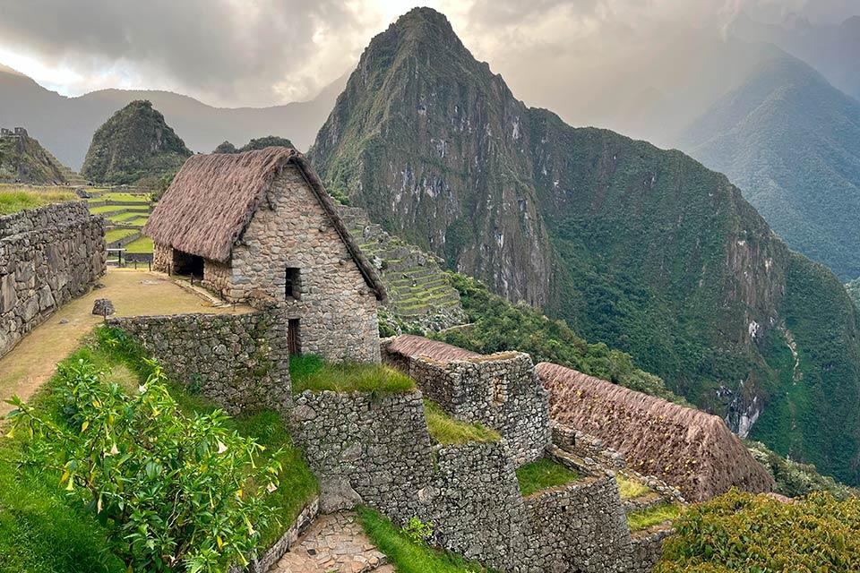 Stone house with a thatched roof in Machu Picchu, with terraces and Huayna Picchu behind under a cloudy sky, Cachicata Inca Quarry Trek to Machu Picchu 4 Days 3 Nights | TreXperience