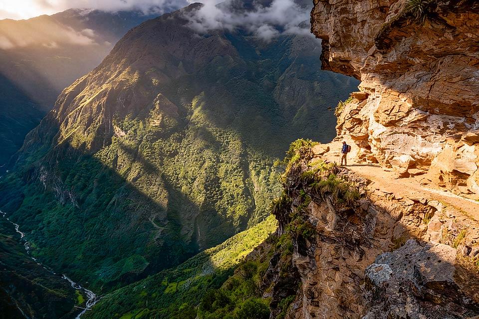 Narrow cliffside trail carved into rock above a deep valley, with a hiker and mountains ahead, Choquequirao Machu Picchu 7 Days Trek | TreXperience