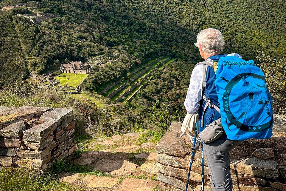 Hiker with a blue backpack looks over Choquequirao terraces and stone buildings from a hillside viewpoint, Choquequirao Machu Picchu 7 Days Trek | TreXperience