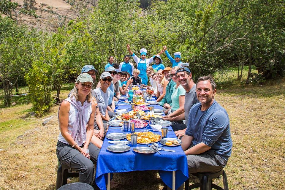 Group seated at a long outdoor table with a blue tablecloth and food served, with the crew in the back raising their hands, Choquequirao Machu Picchu 7 Days Trek | TreXperience