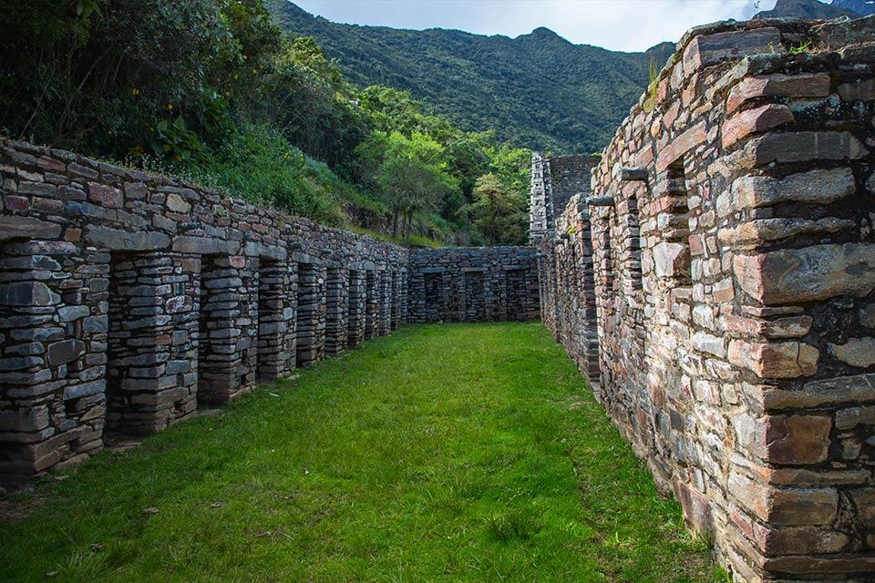 Pasaje entre muros de piedra con puertas bajas y césped al centro, con ladera verde y montañas al fondo, Choquequirao Trek 4 días 3 noches | TreXperience