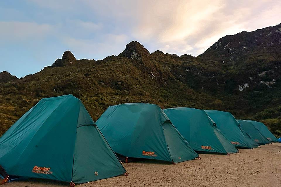 Fila de carpas verdes instaladas en un campamento sobre suelo de tierra, con montañas oscuras al fondo, Choquequirao Trek 4 días 3 noches | TreXperience